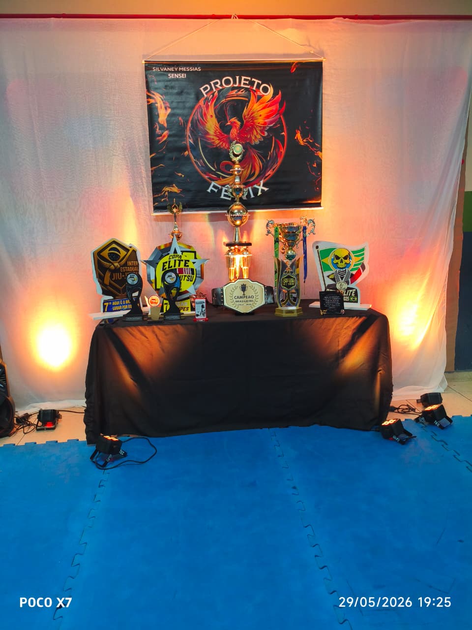 Mesa com troféus e cinturão sob o banner do Projeto Fênix