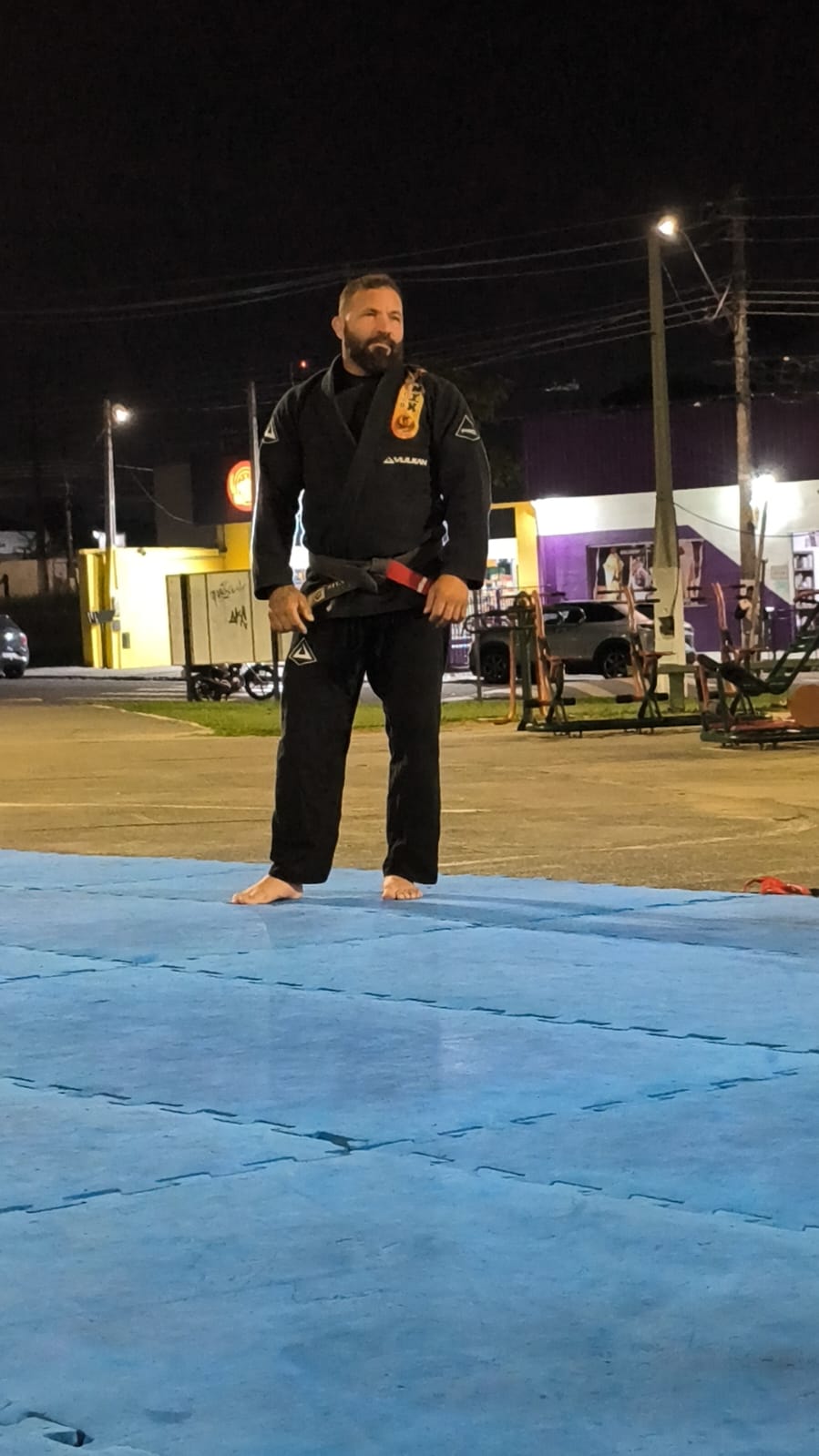Faixa preta em pé no tatame ao ar livre durante treino noturno