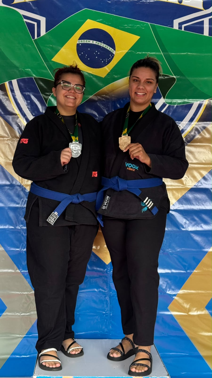 Duas alunas adultas faixa azul com medalhas ao lado da bandeira do Brasil